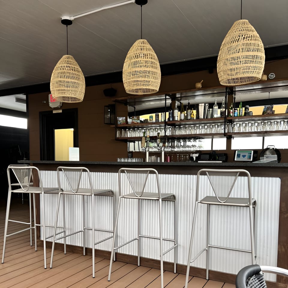 An empty bar with four stools, wicker pendant lights, and shelves of bottles and glasses.