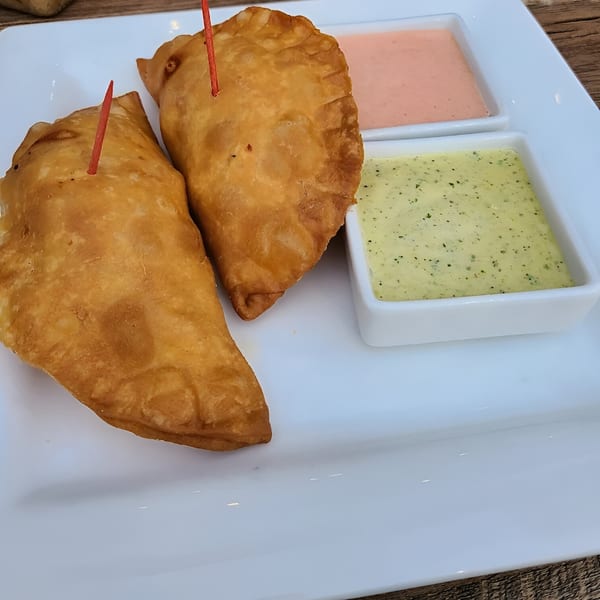 Two golden-brown empanadas with red toothpicks, served with pink and green dipping sauces on a white plate.