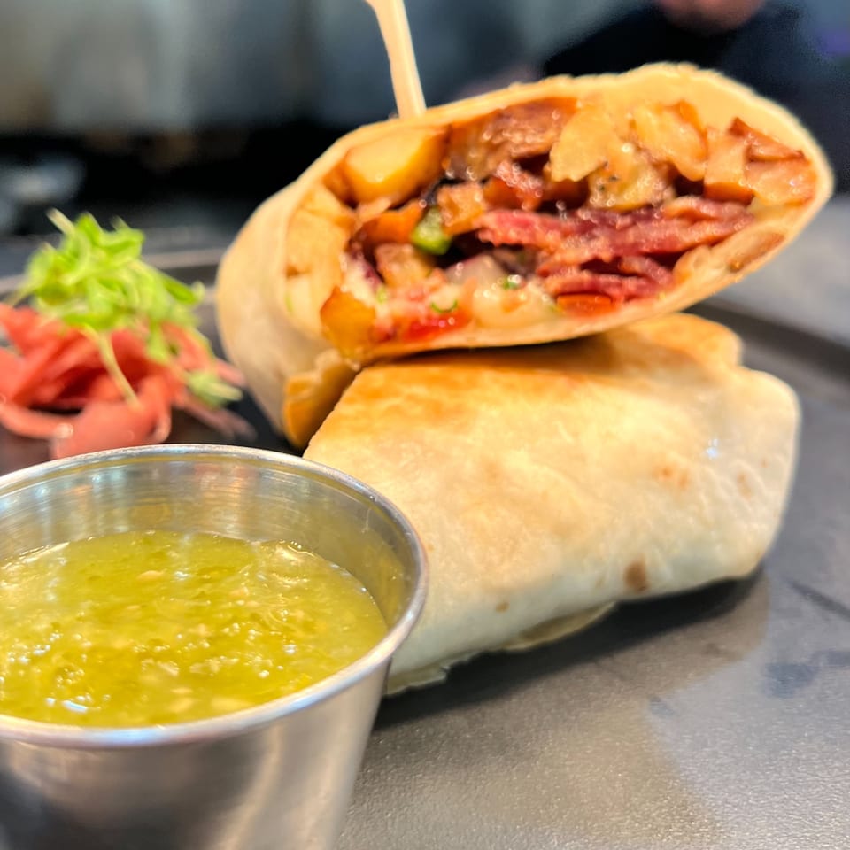 A breakfast burrito, cut in half, revealing a filling of meat and potatoes, served with green salsa.